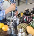 Hands mixing ingredients in a metal cocktail shaker beside fruit, glasses and bottles.