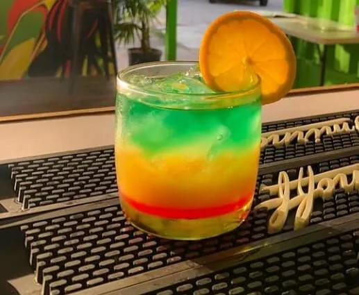 A layered red, yellow and green cocktail with ice and an orange-slice garnish on a bar mat.